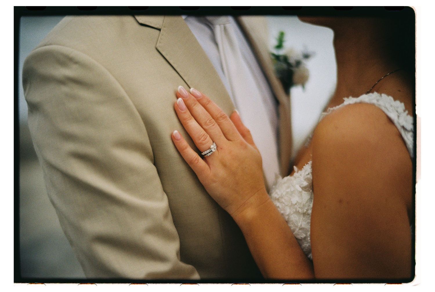 A close-up shot shows a hand wearing a wedding ring pressed against a beige suit jacket at a wedding ceremony.