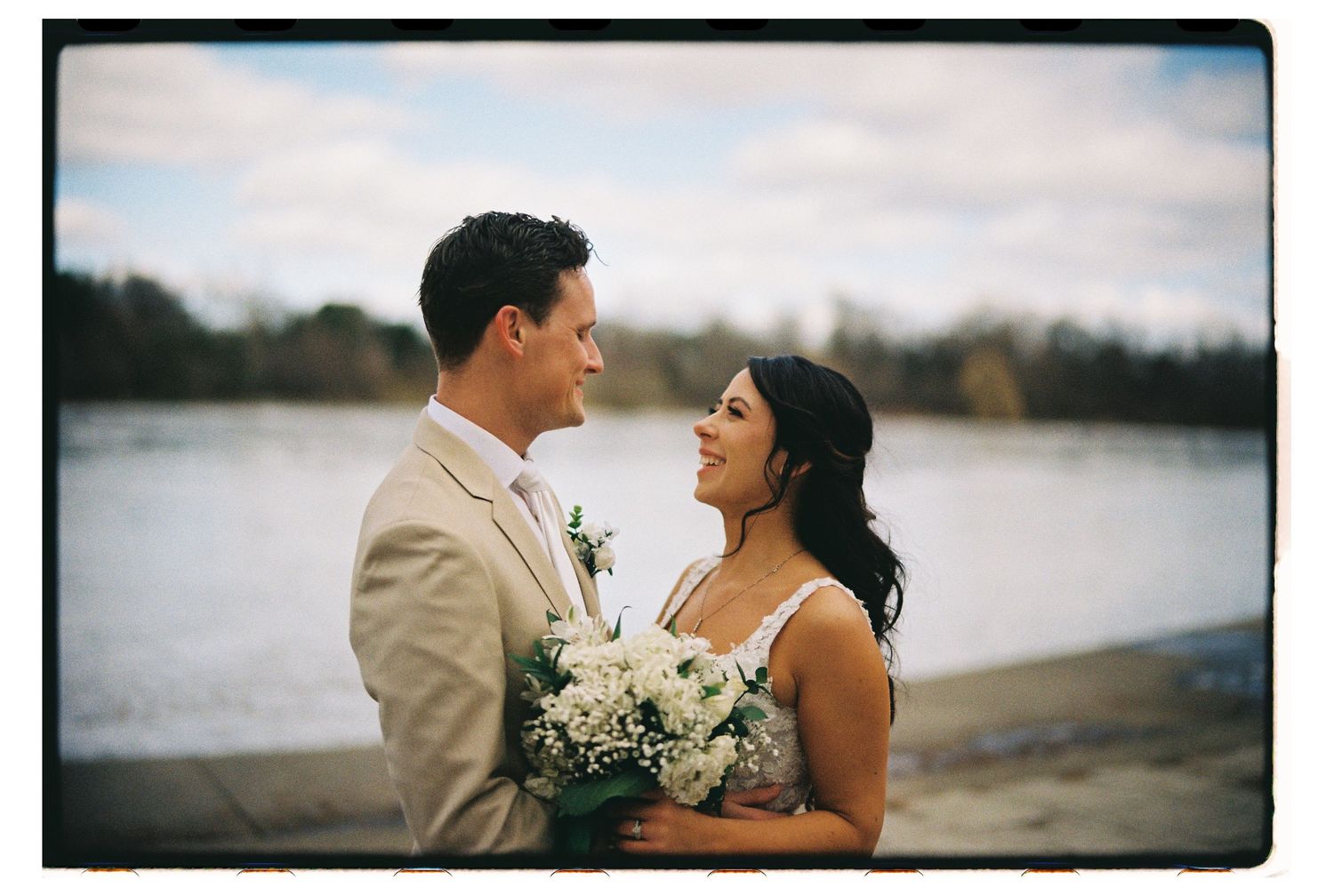 A couple in wedding attire embrace and smile at each other while holding a white bouquet by a lakeside.