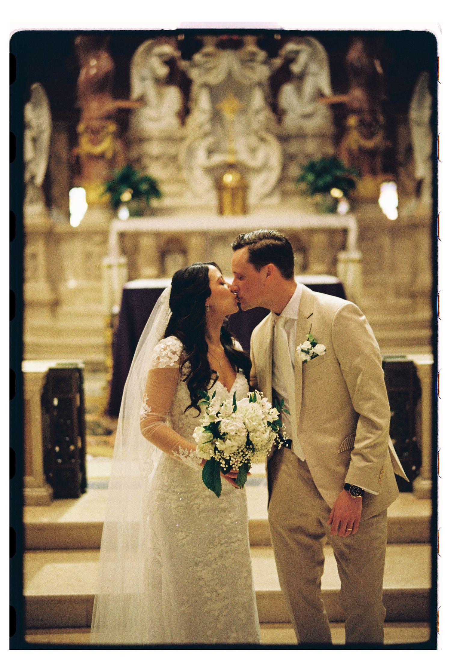 Newlyweds share a romantic kiss at the altar of an ornate church with white flowers and a tan suit.