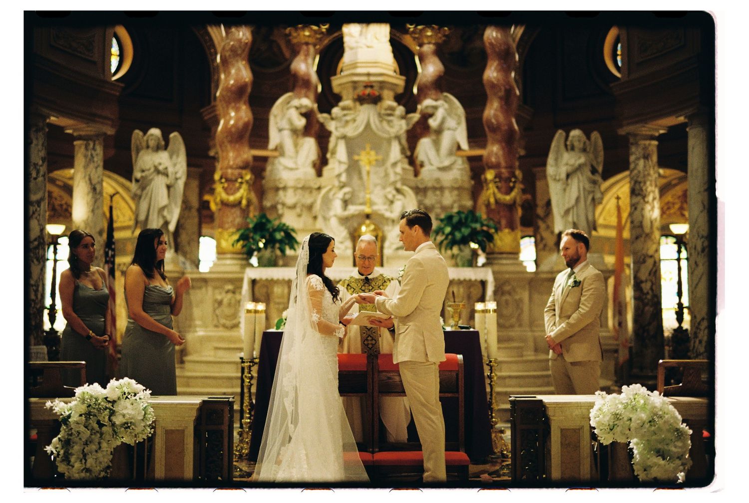 Traditional wedding ceremony taking place in an ornate Catholic church with gilded altar and white floral arrangements.