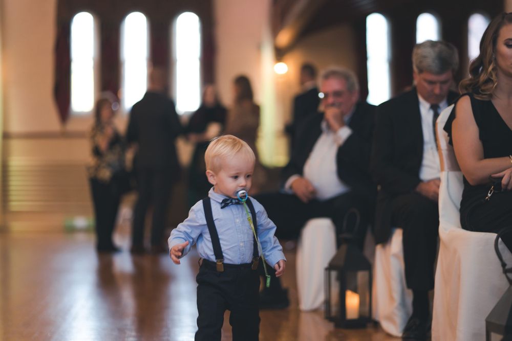 A young child in suspenders walks across a wooden dance floor at a formal event.