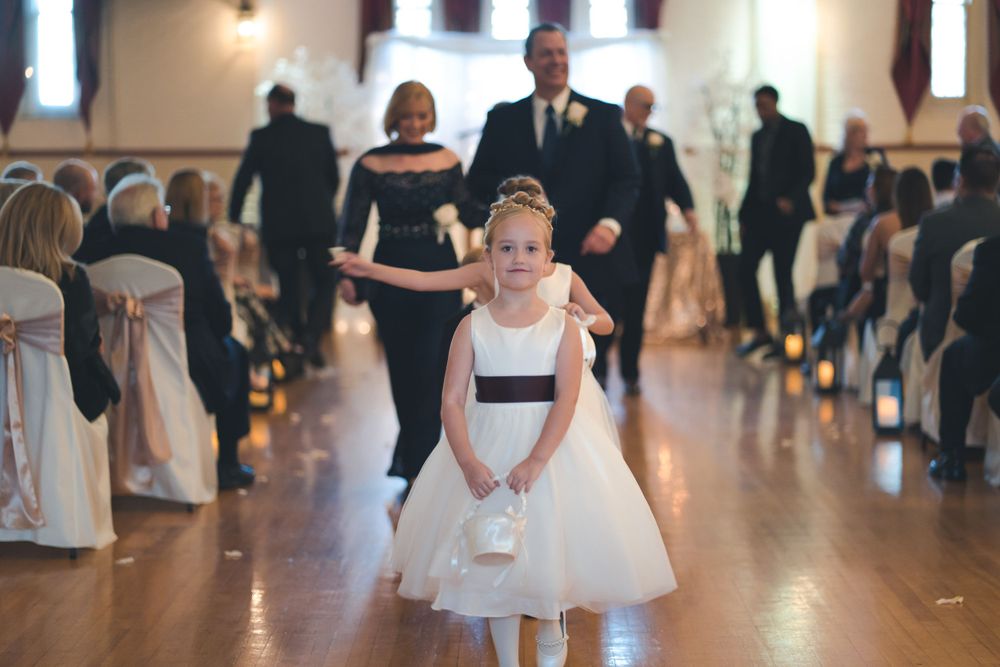 A flower girl in a white tulle dress walks down the aisle at a formal wedding ceremony.