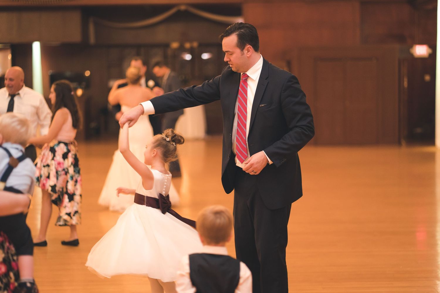 A man in a suit gestures on the dance floor near a child in a white dress.