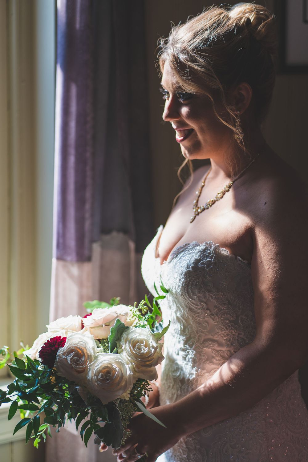 Bride in lace gown holding rose bouquet by window in natural light.