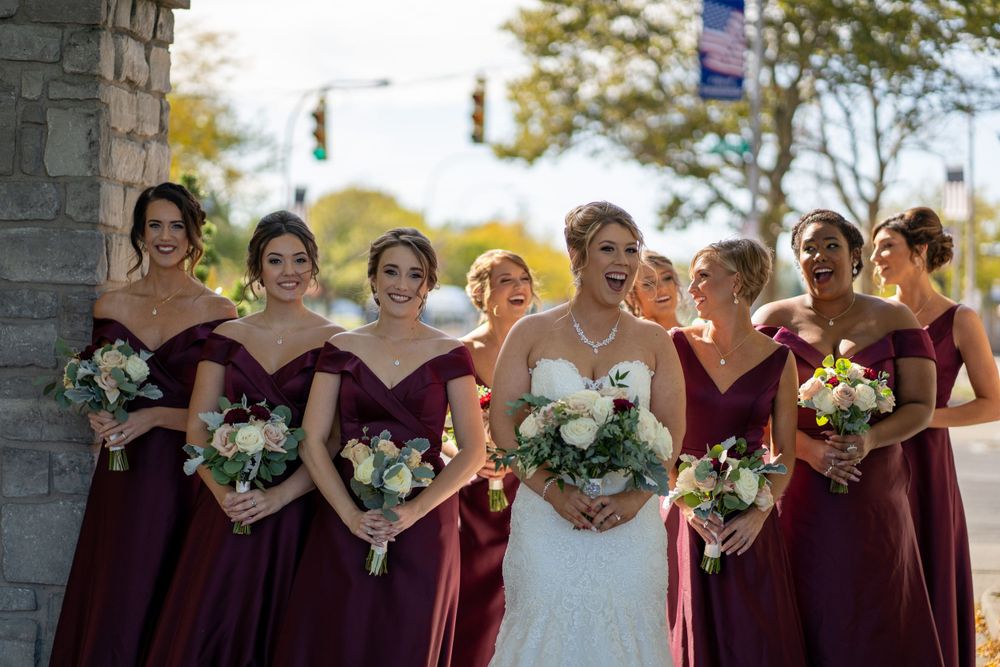 A wedding party with bridesmaids in burgundy dresses holding white floral bouquets outdoors.