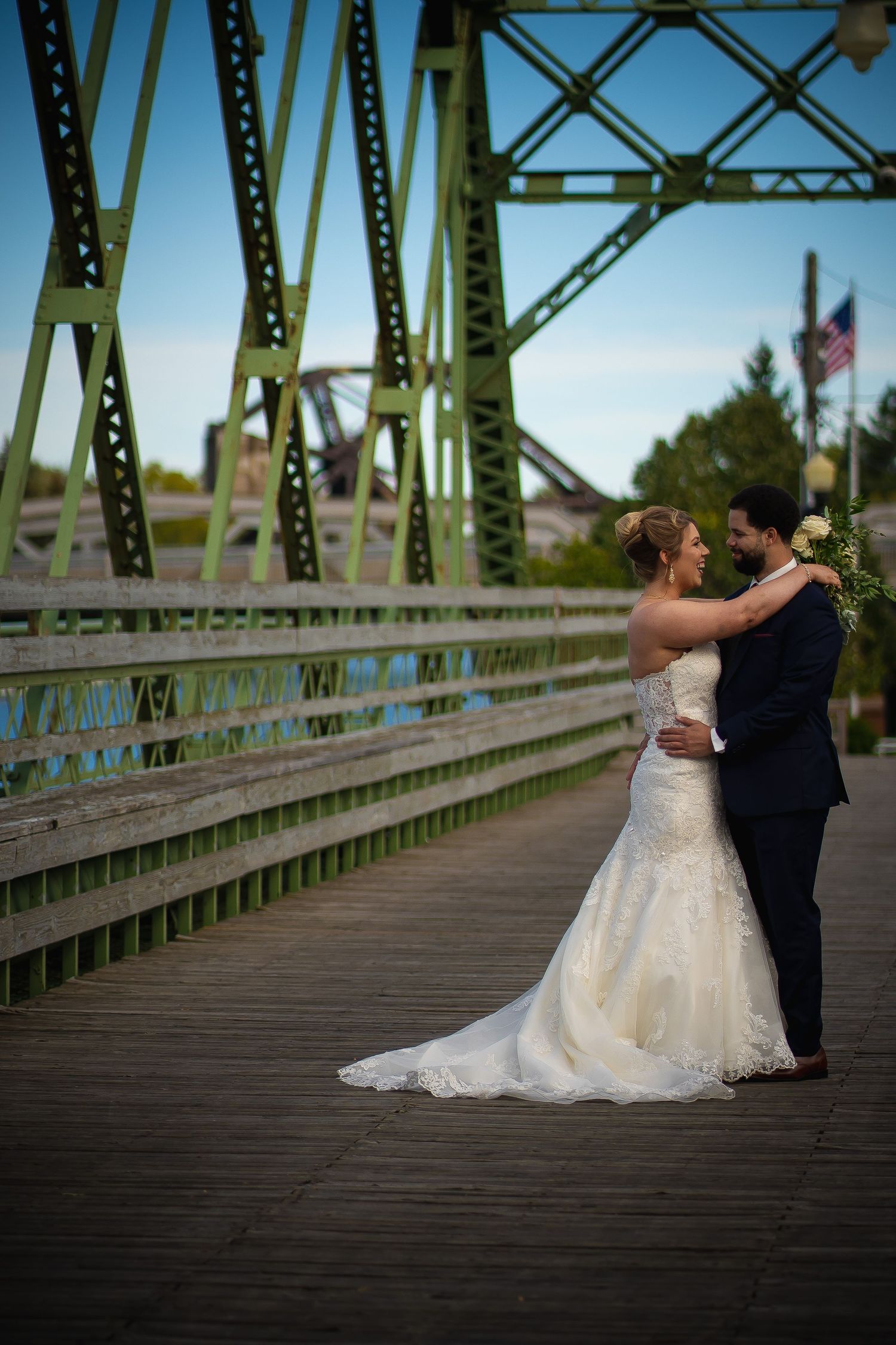 Newlyweds embrace on wooden bridge with green metal structure in background.