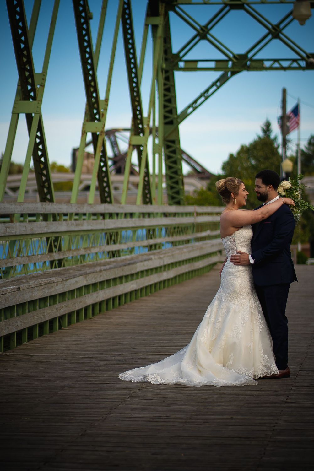 Newlyweds embrace on wooden bridge with green metal structure in background.