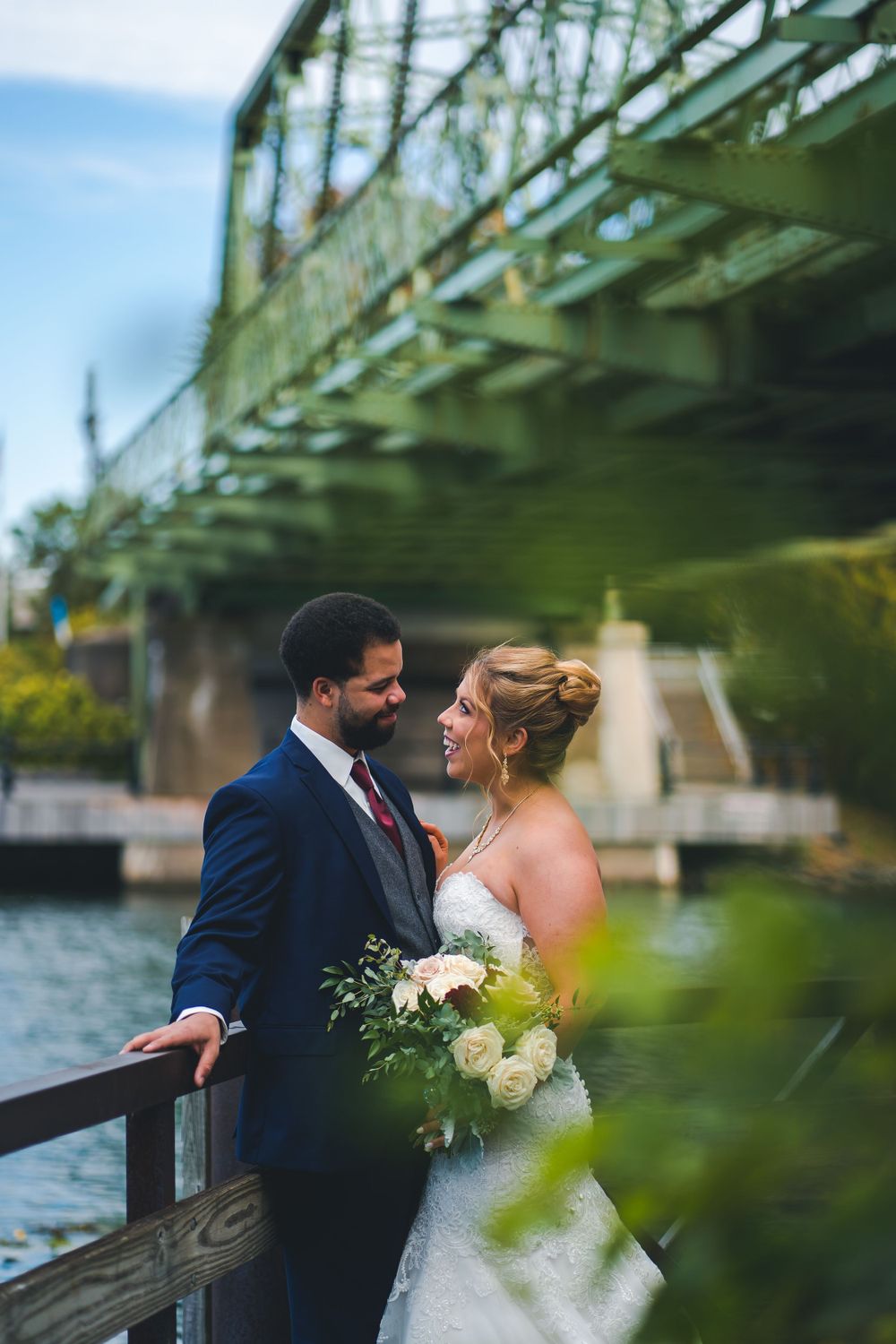 A wedding couple shares an intimate moment by a green metal bridge over water.