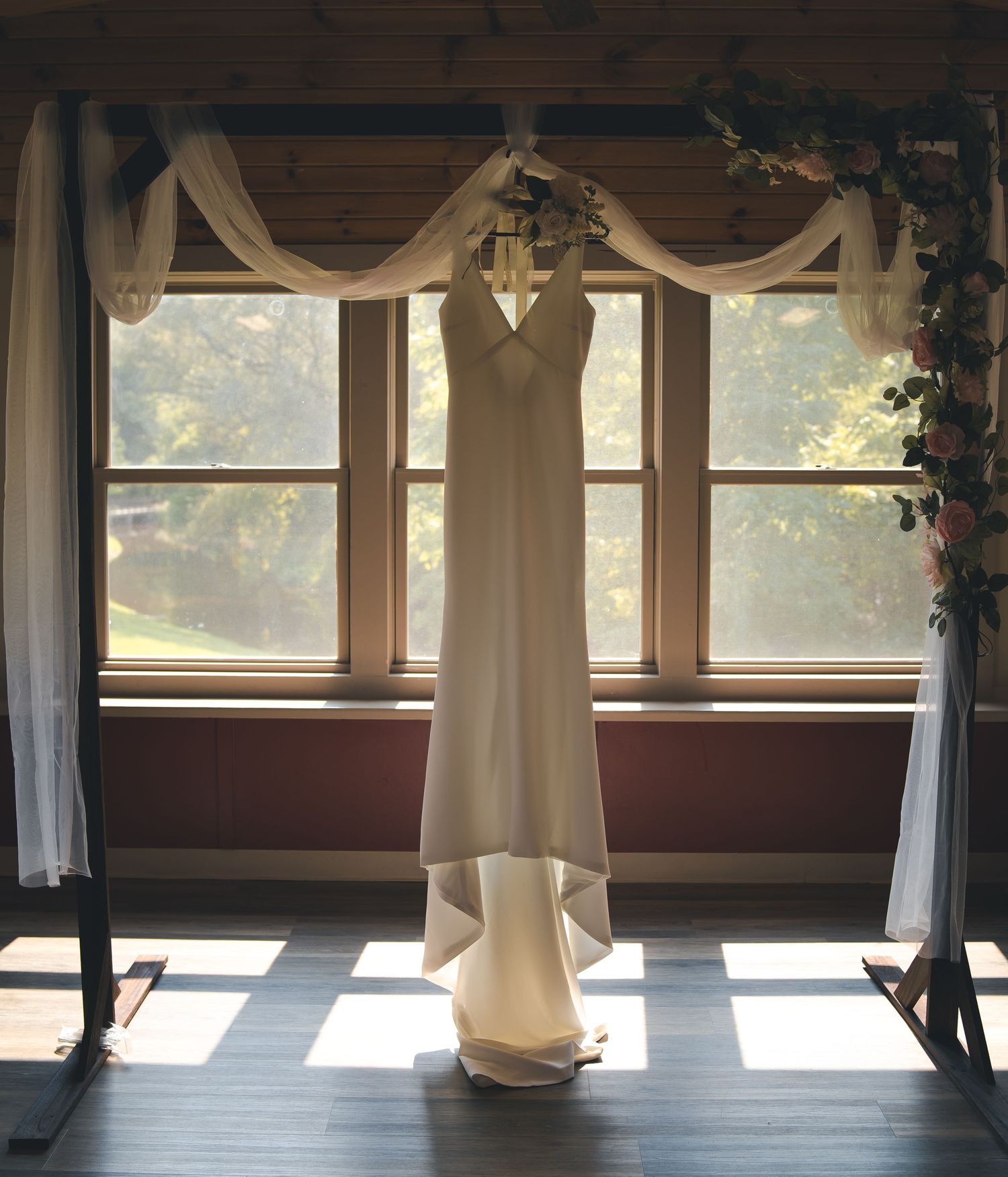 An elegant white wedding dress hangs in front of windows with draped fabric and natural light.