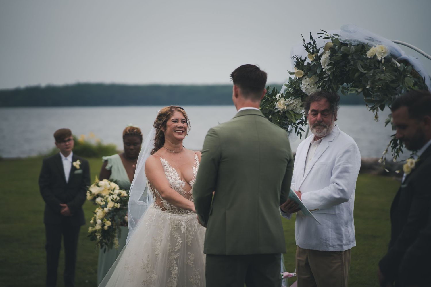 Outdoor waterfront wedding ceremony with formal attire and floral decorations.