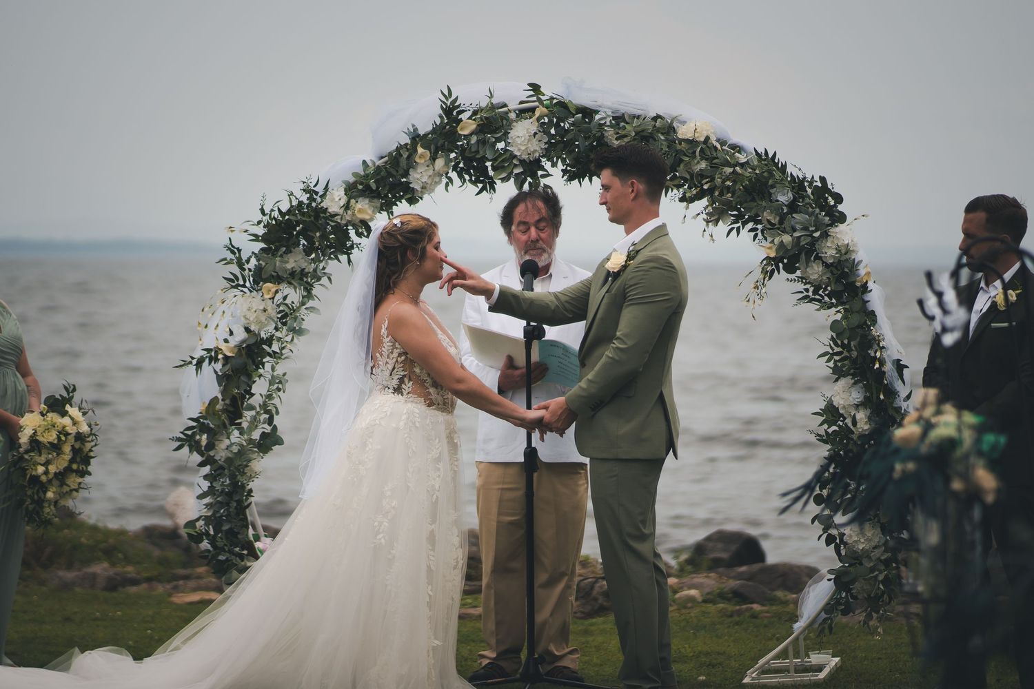 A wedding ceremony takes place under a floral arch overlooking a waterfront with an officiant and couple at sunset.