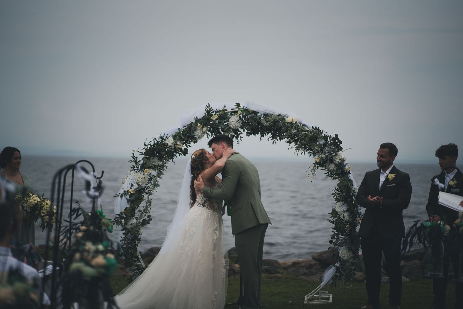 Intimate waterfront wedding ceremony under a greenery arch with guests looking on.
