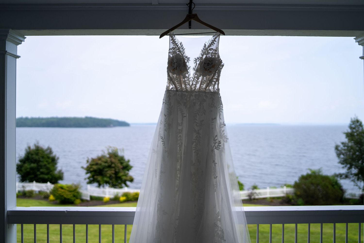 Delicate lace wedding dress hanging in front of large windows with a waterfront view.