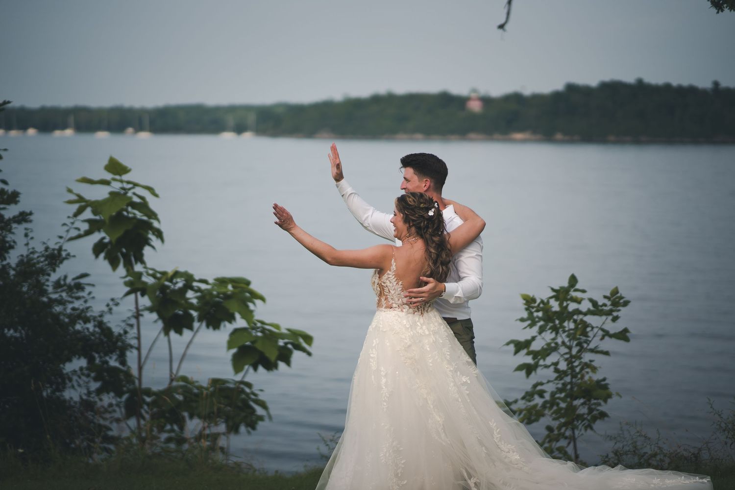 Newlyweds dancing by a lakeside in wedding attire.