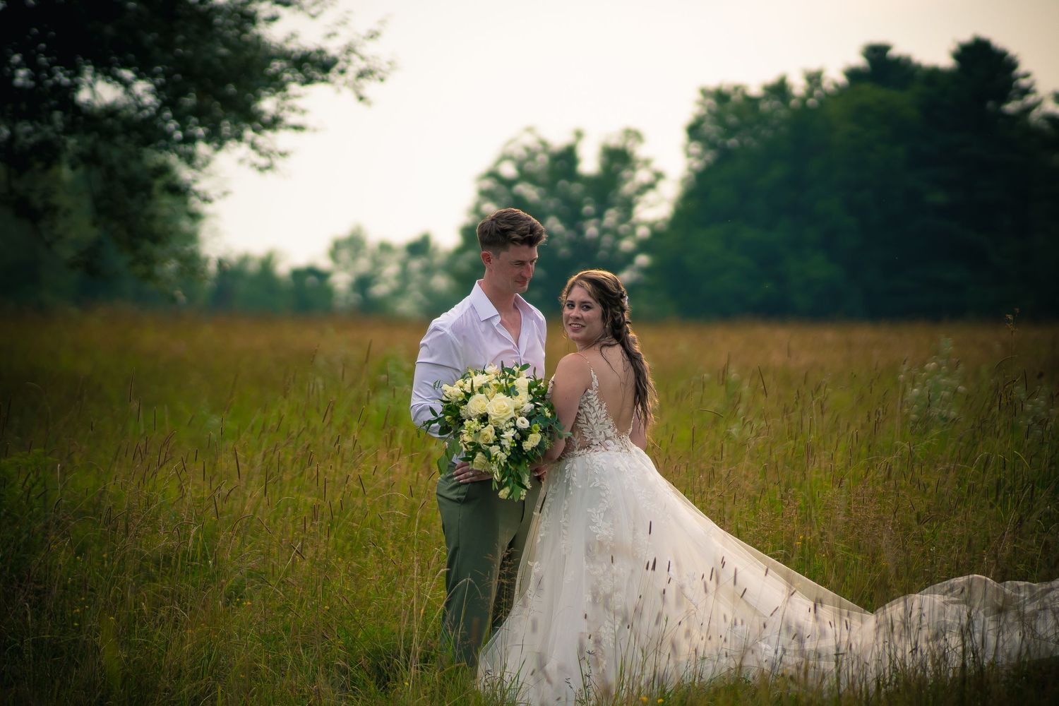 Wedding couple poses in golden summer field with bouquet at sunset.