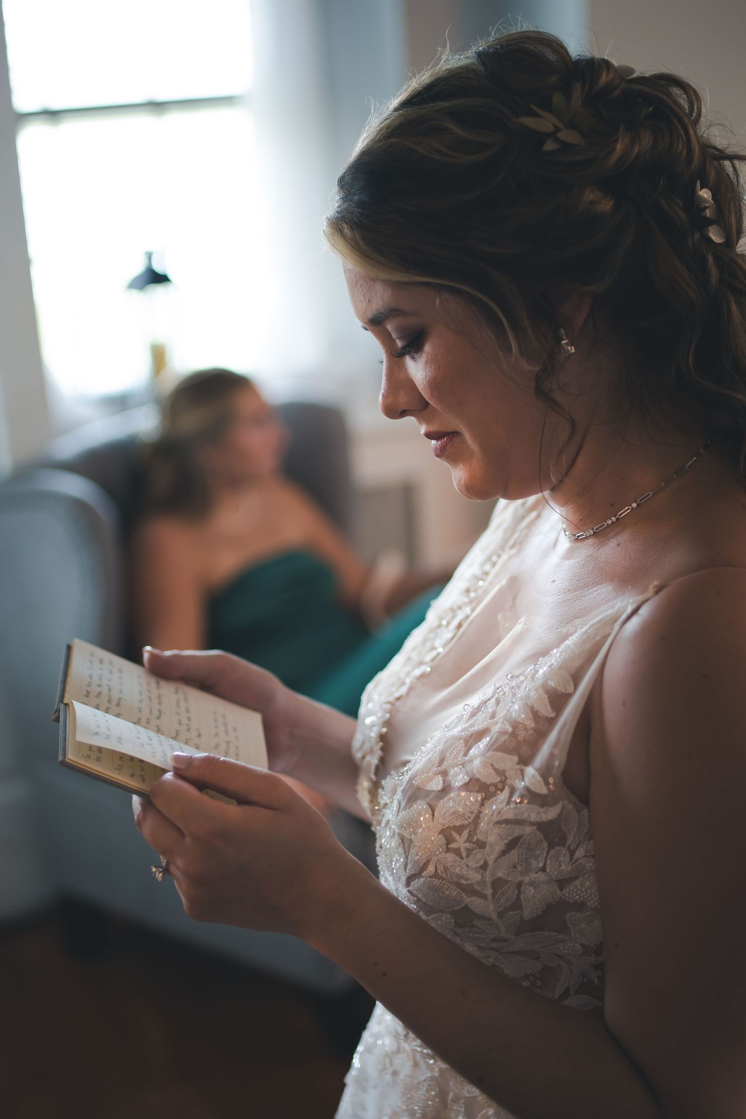 A bride in a lace wedding gown reads from a small book during wedding preparations.