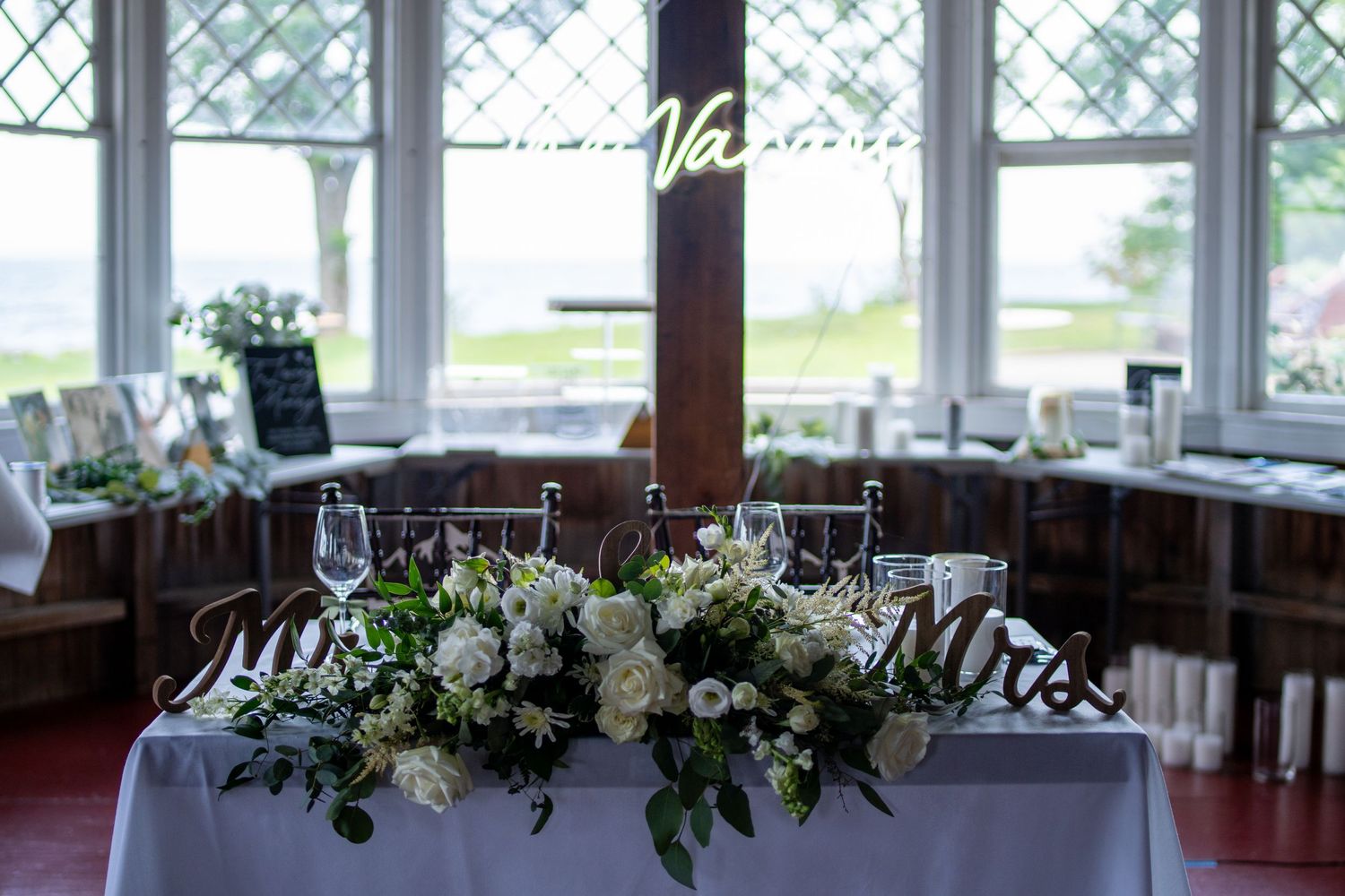 White floral centerpiece arrangement on a table with wine glasses and a waterfront view through windows.