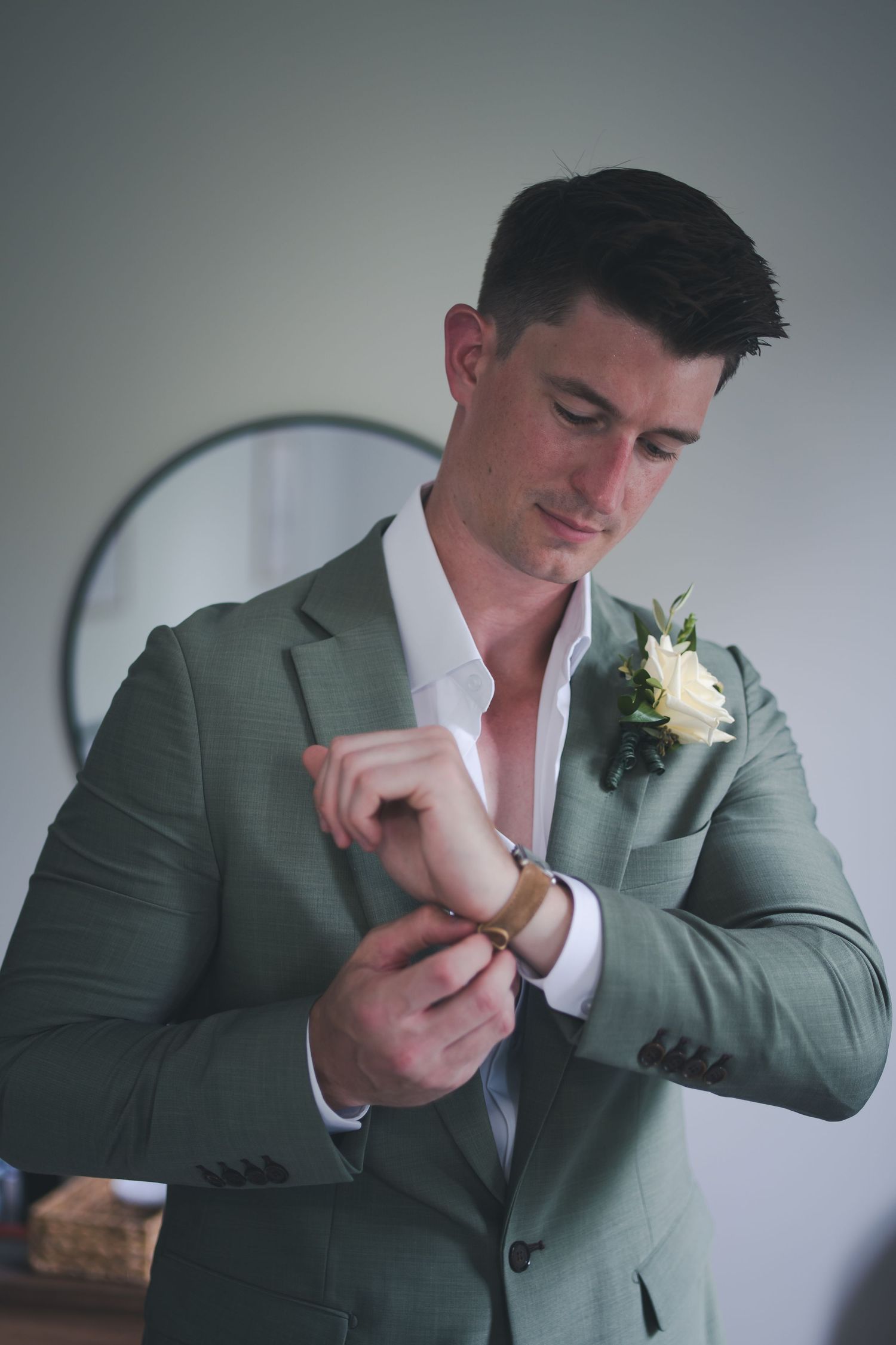 A groom in sage green suit adjusts his cufflinks while getting ready.