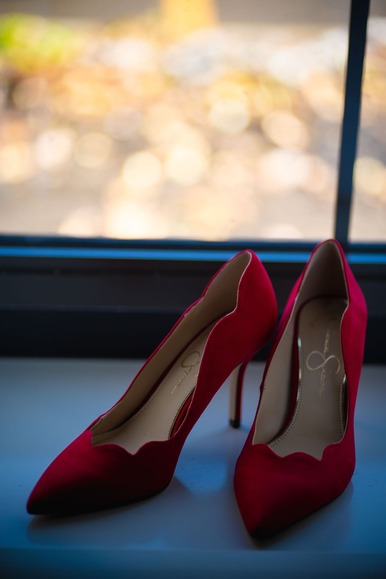 A pair of elegant red suede high heel pumps sits on a windowsill with blurred background.