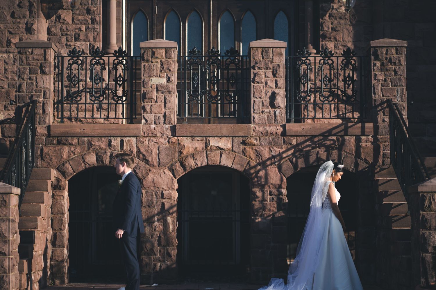 A wedding couple poses against a gothic stone building with arched windows.