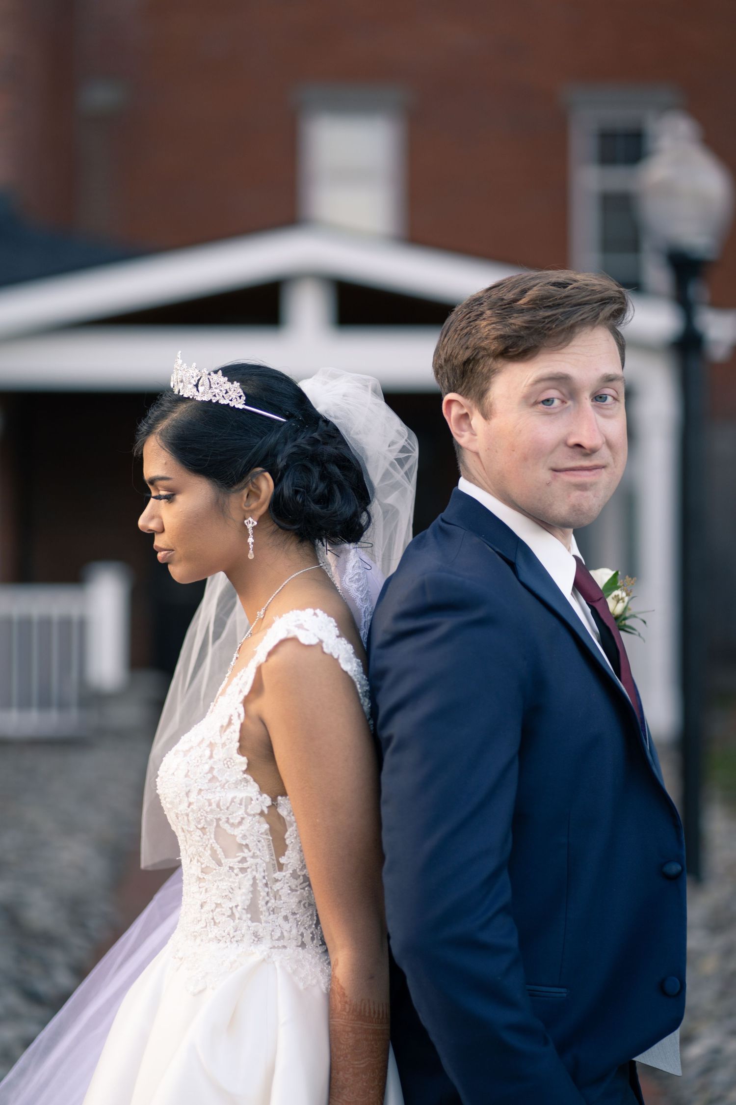 A bride in a lace gown and veil stands back-to-back with groom in navy suit.
