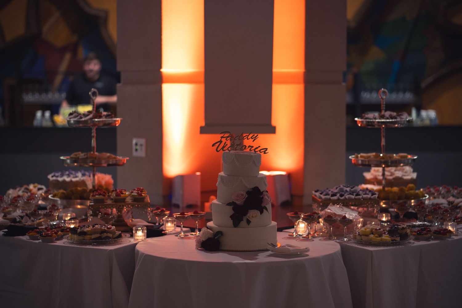 Elegant wedding cake display with desserts on a table illuminated by warm orange lighting.