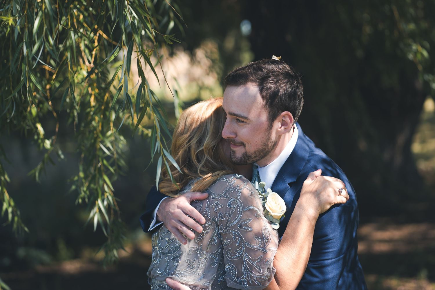 A tender embrace between newlyweds beneath willow trees in a romantic outdoor setting.