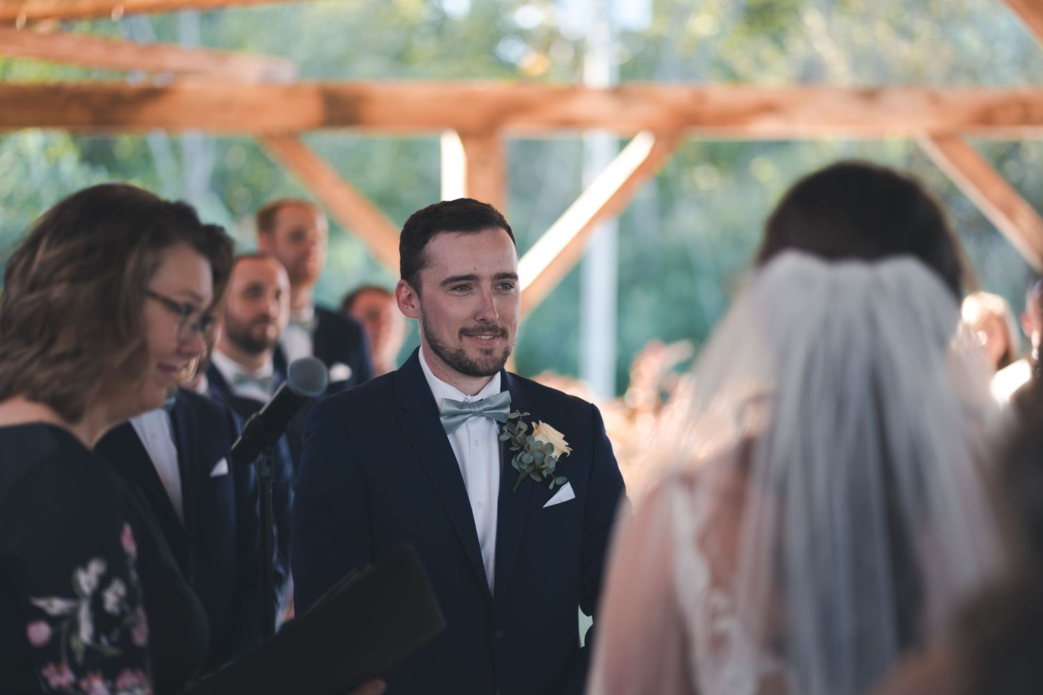 A wedding ceremony moment with guests gathered under a rustic wooden pergola.