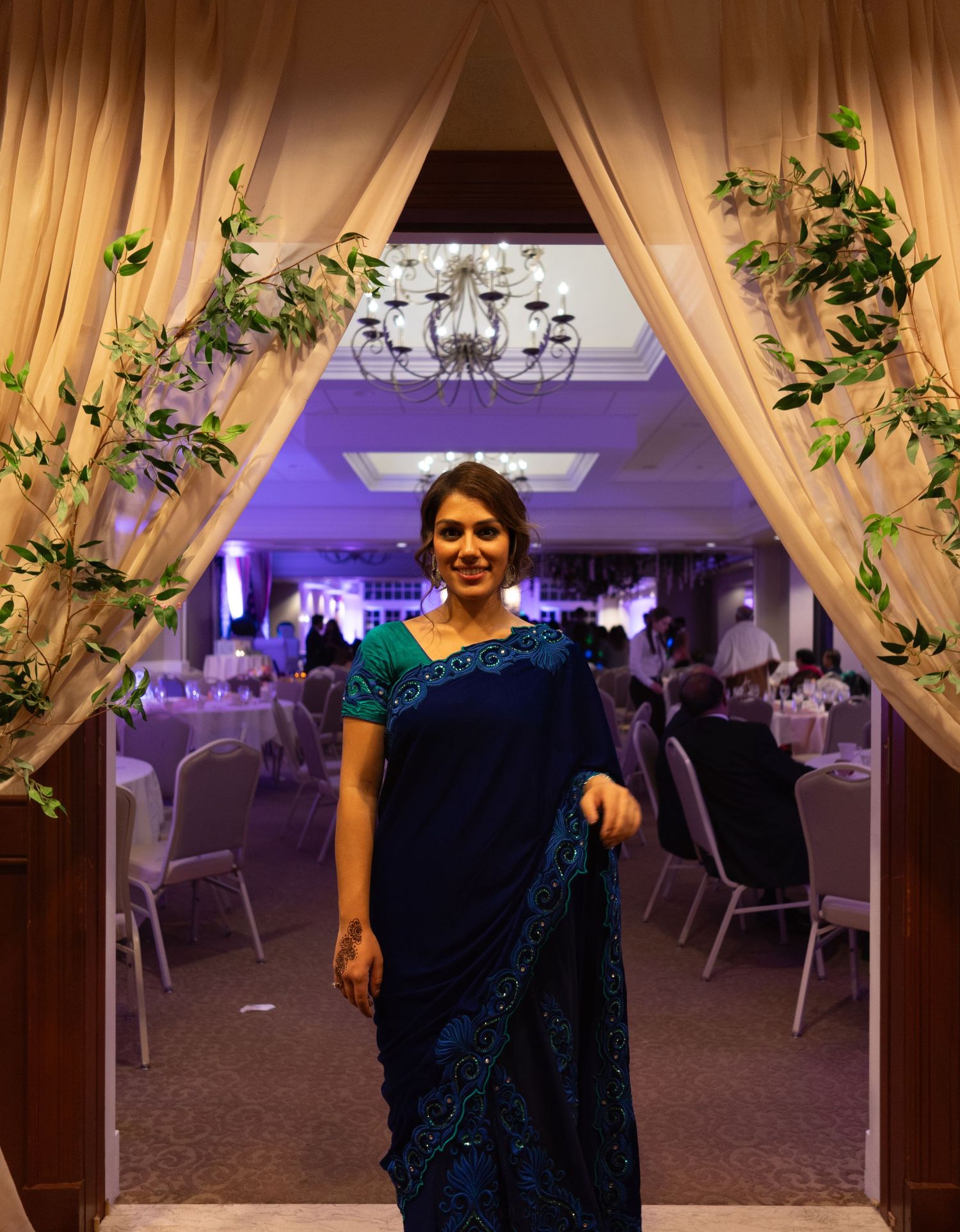 An elegant evening event setup with cream curtains, purple uplighting and floral decor in a banquet hall.