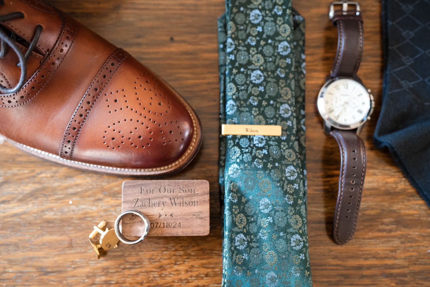 Groom's accessories featuring brown leather shoes, watch, tie and wedding rings on wooden surface.