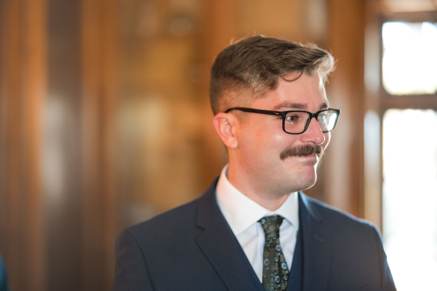 A formal portrait in a warmly lit hallway featuring a navy suit and glasses.