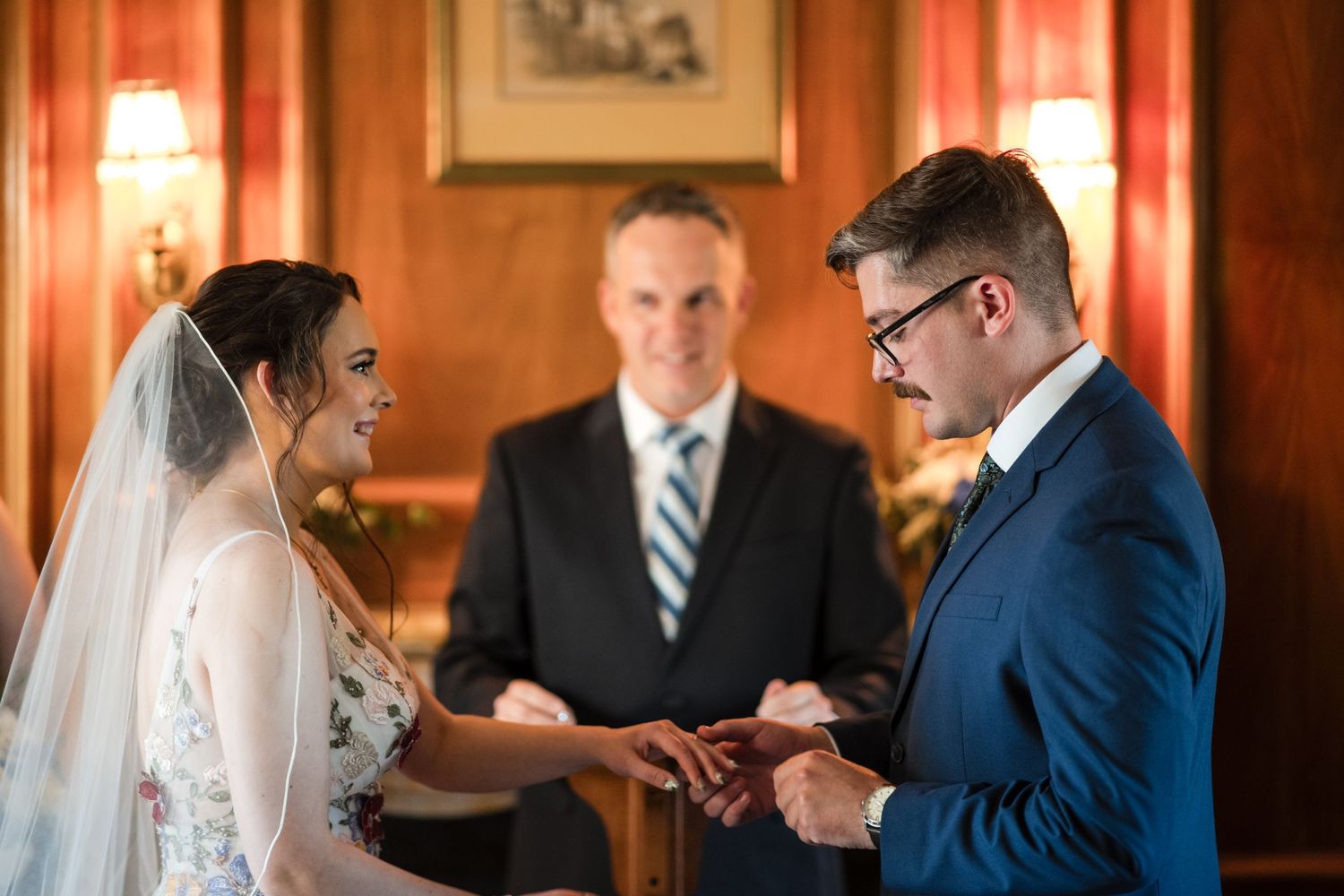 Exchanging rings during an indoor wedding ceremony with wood paneling and soft lighting.