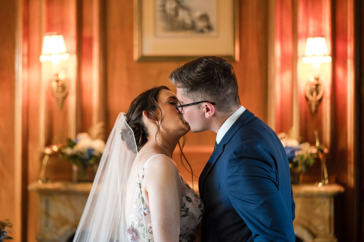 A romantic wedding kiss in an ornate room with wall sconces and warm ambient lighting.