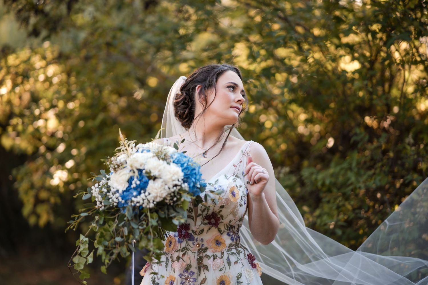Bride in floral dress holds blue and white bouquet against golden autumn backdrop.