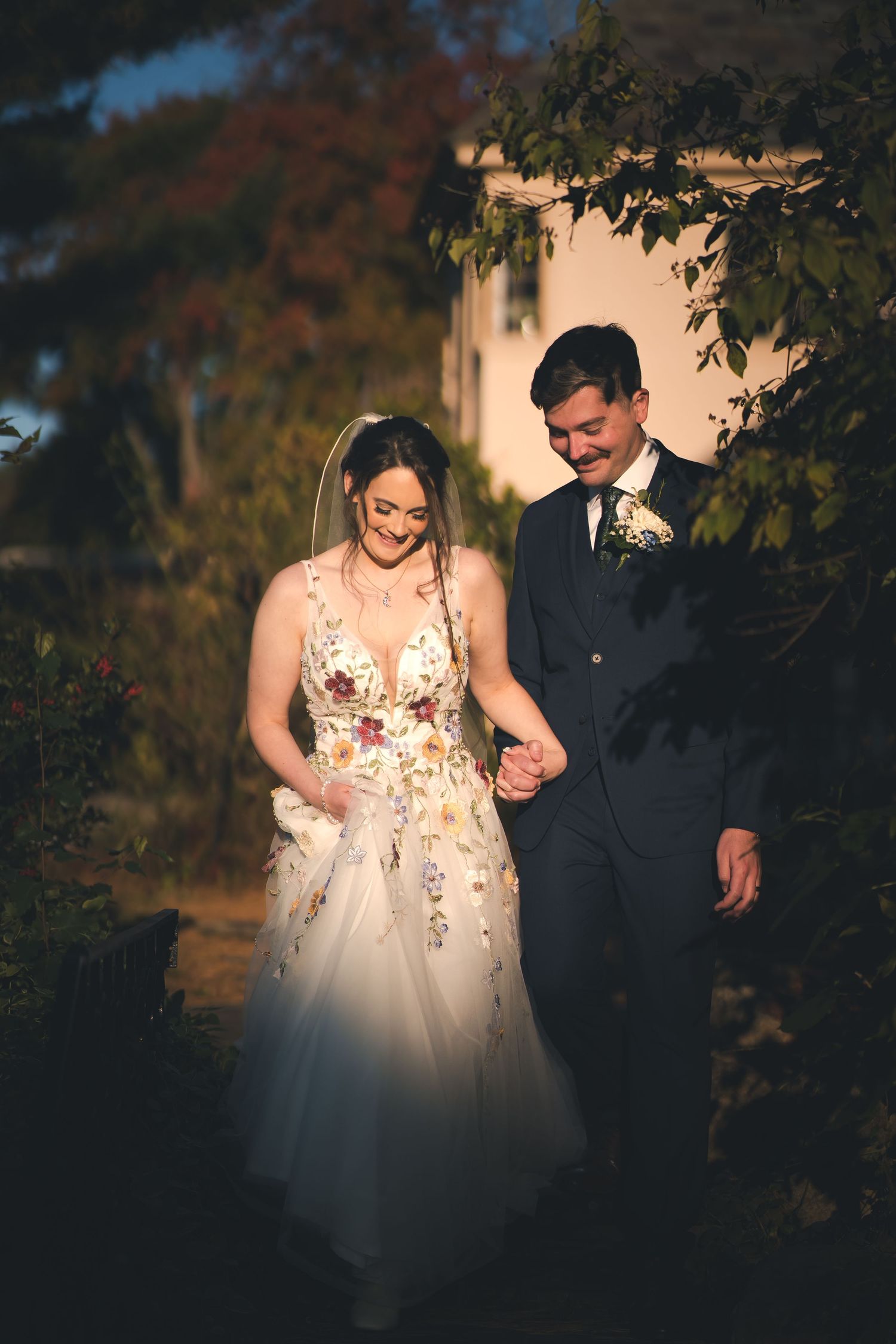 A couple walks together at dusk, bride wearing a floral wedding dress against autumn foliage.
