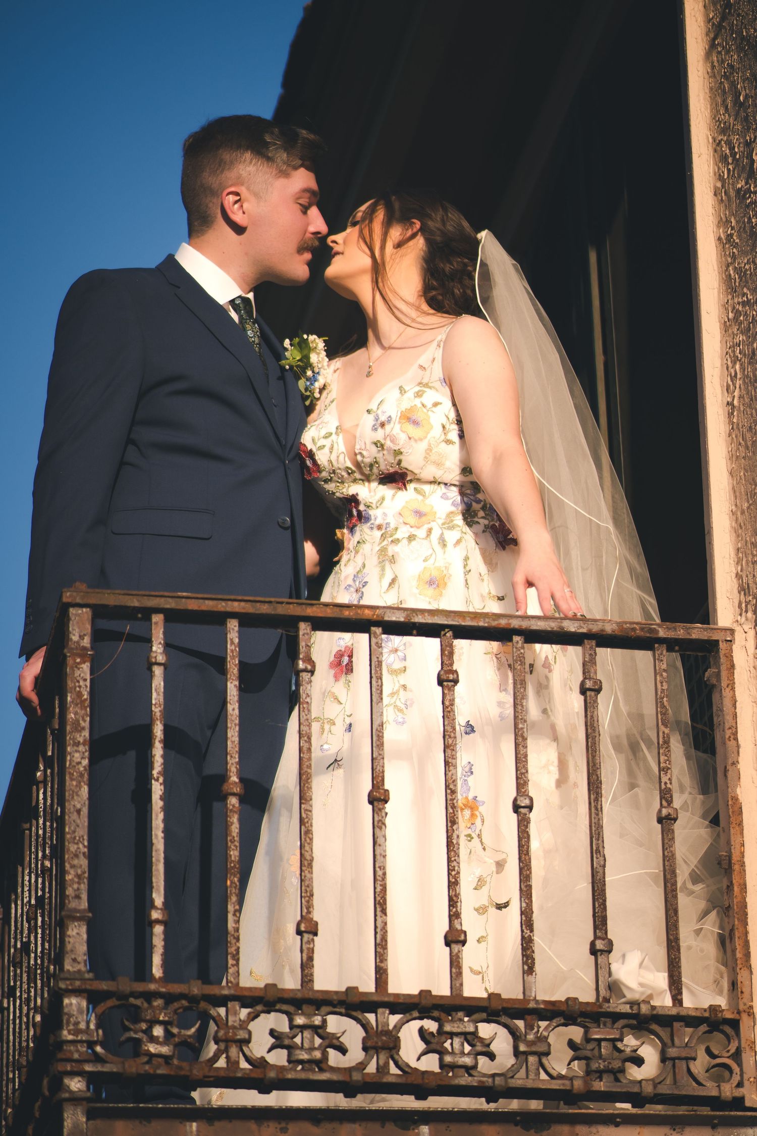 Newlyweds share an intimate moment on an ornate balcony against blue sky.