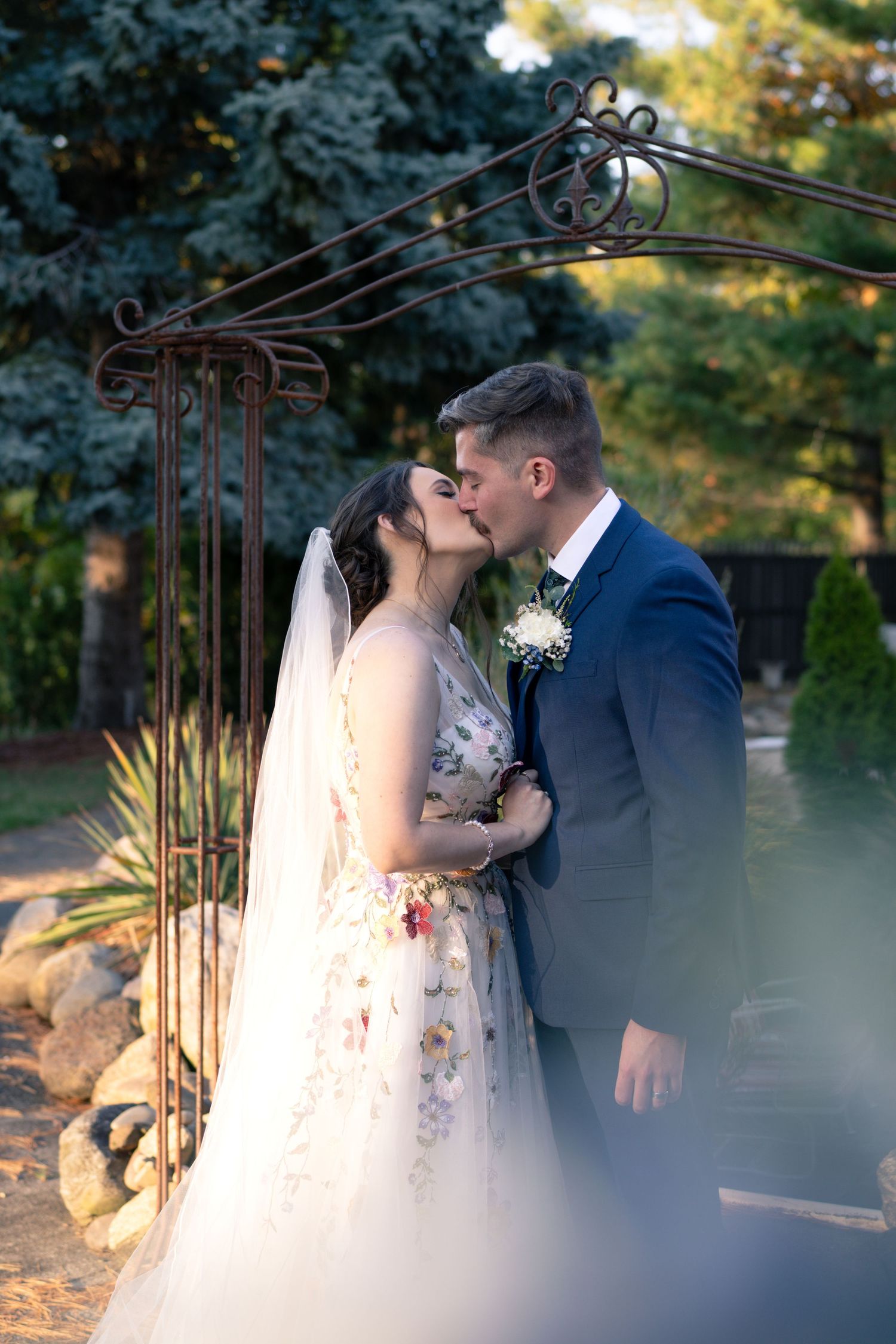 Romantic wedding kiss under an ornate garden archway with evergreens in background.