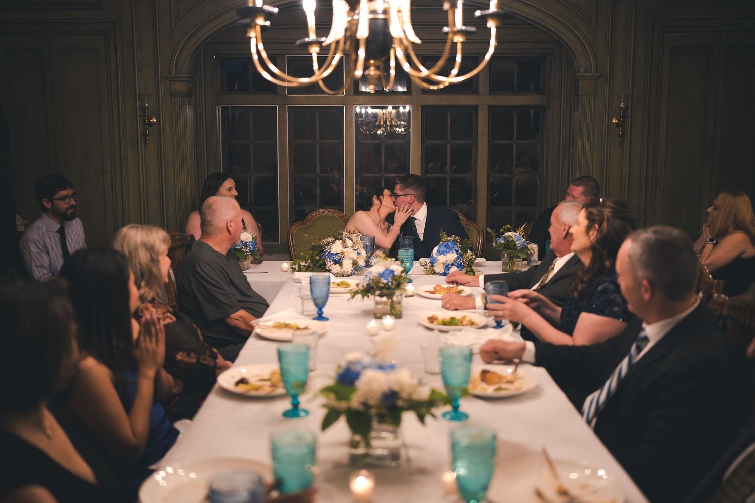 Intimate wedding reception dinner under a chandelier with guests seated at a long table.
