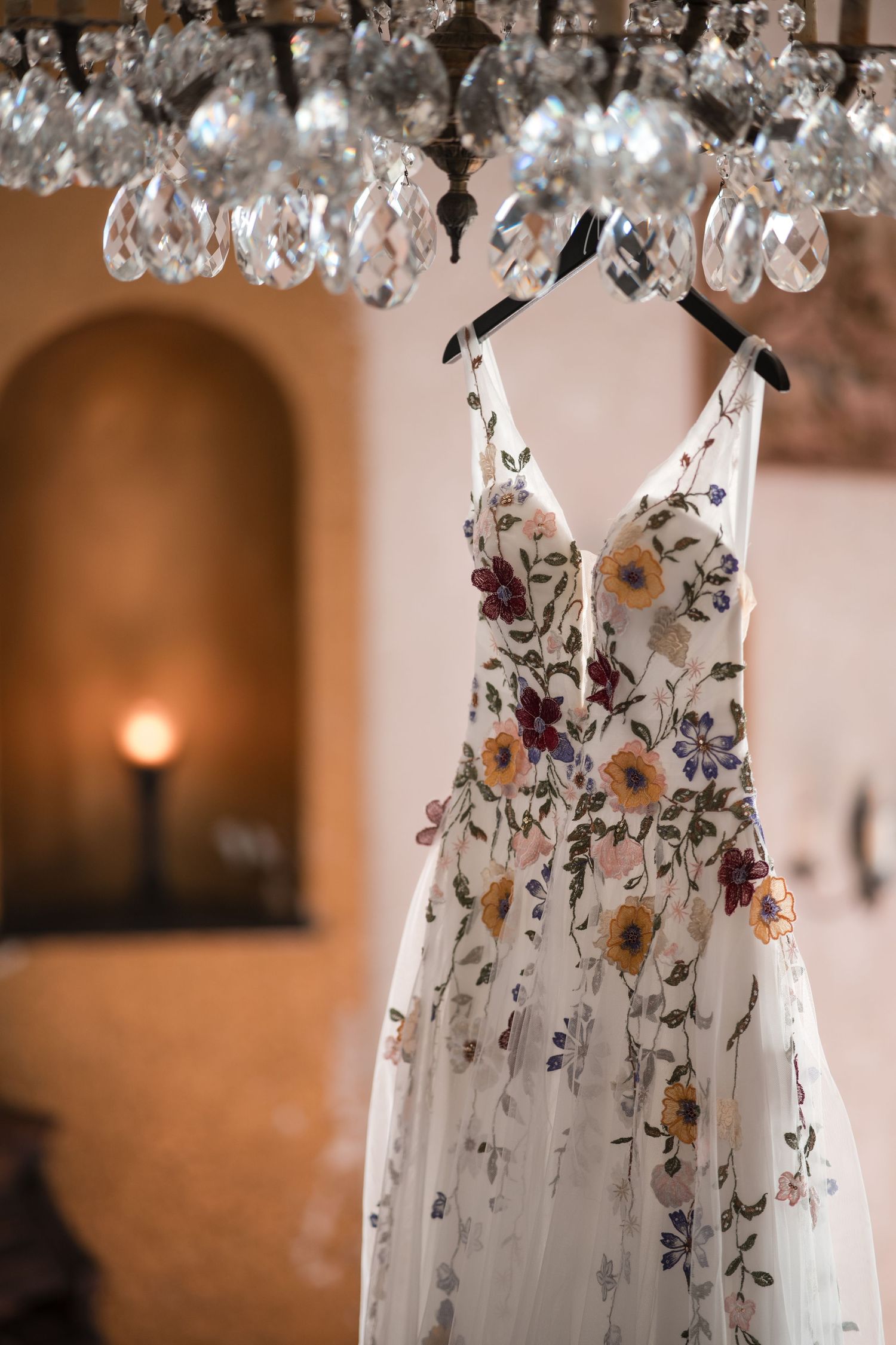 Floral wedding dress with embroidered flowers hanging from crystal chandelier in warm lighting.