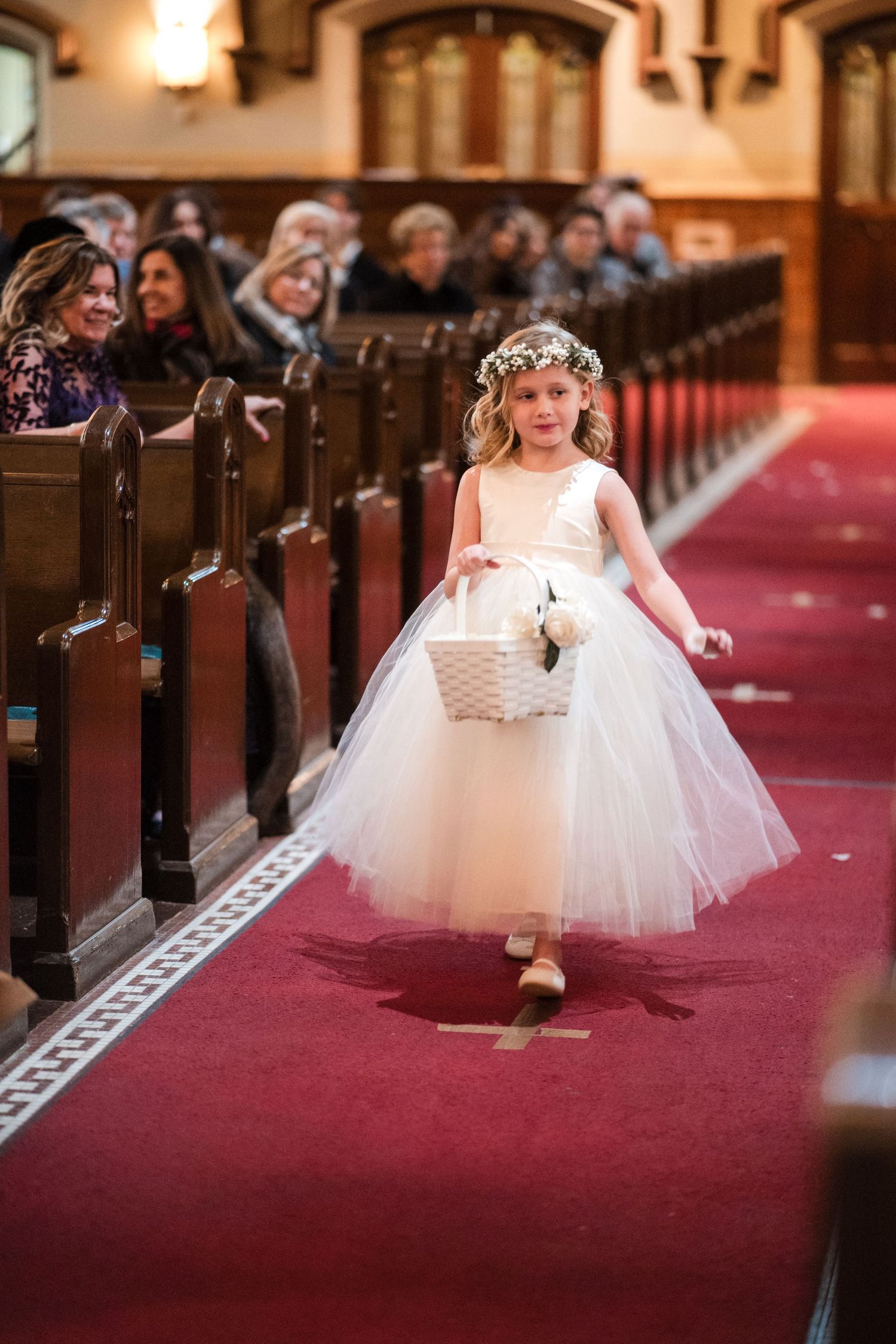 A young flower girl in a white tulle dress walks down the red-carpeted church aisle carrying a basket during a wedding ceremony.