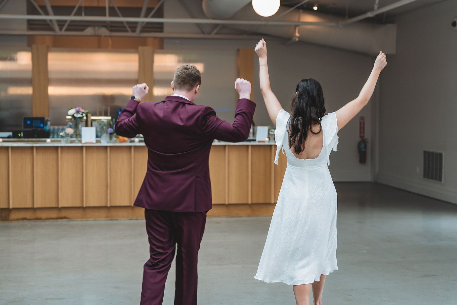 Two people dancing together in a spacious room, one wearing a burgundy suit and one in a flowy white dress.