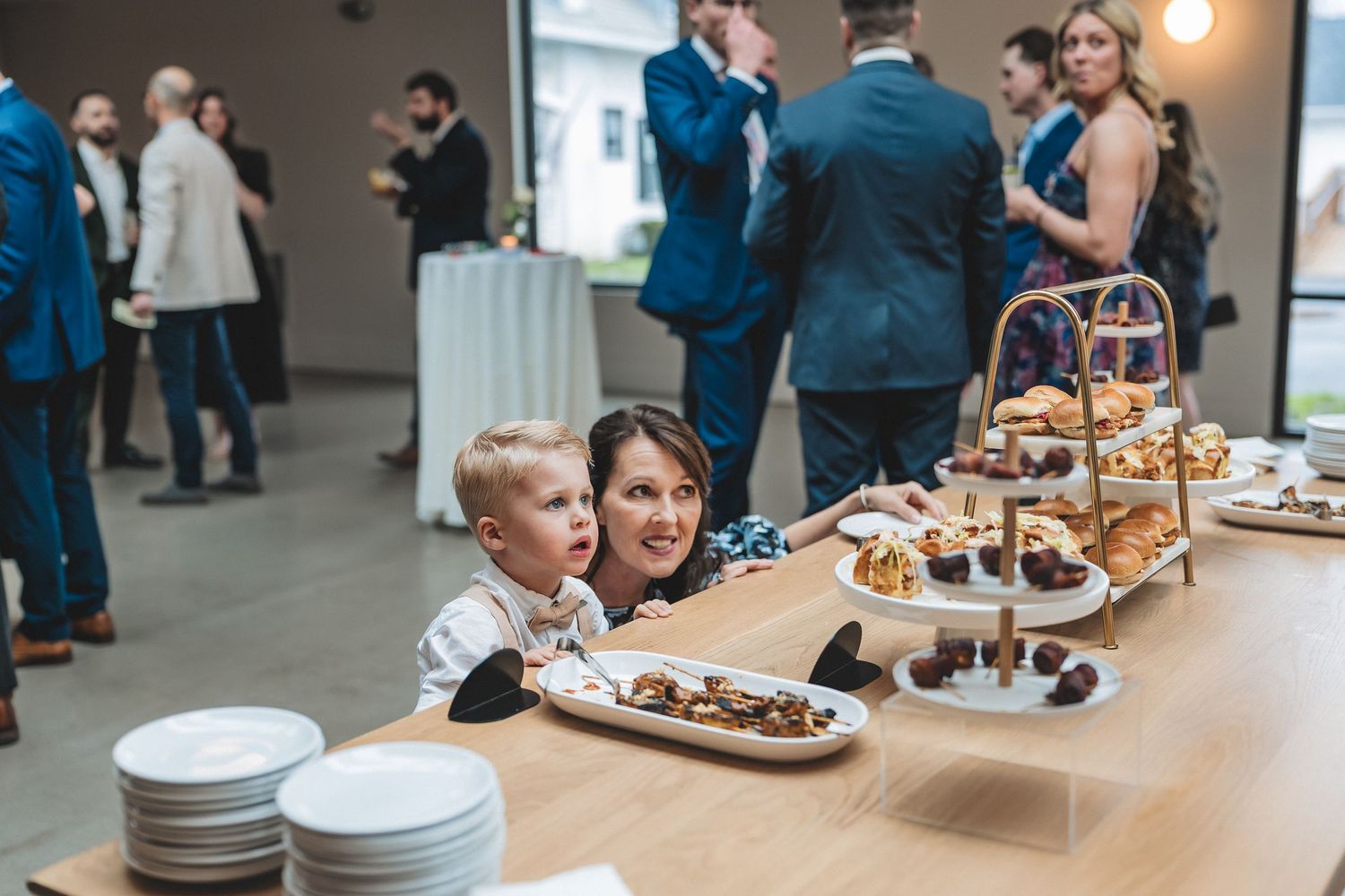 People gather around a dessert display table at an event with a tiered serving stand filled with treats.