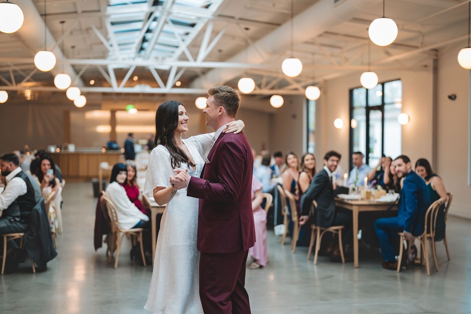 A couple shares their first dance in a modern venue with round pendant lights and guests seated at tables in the background.