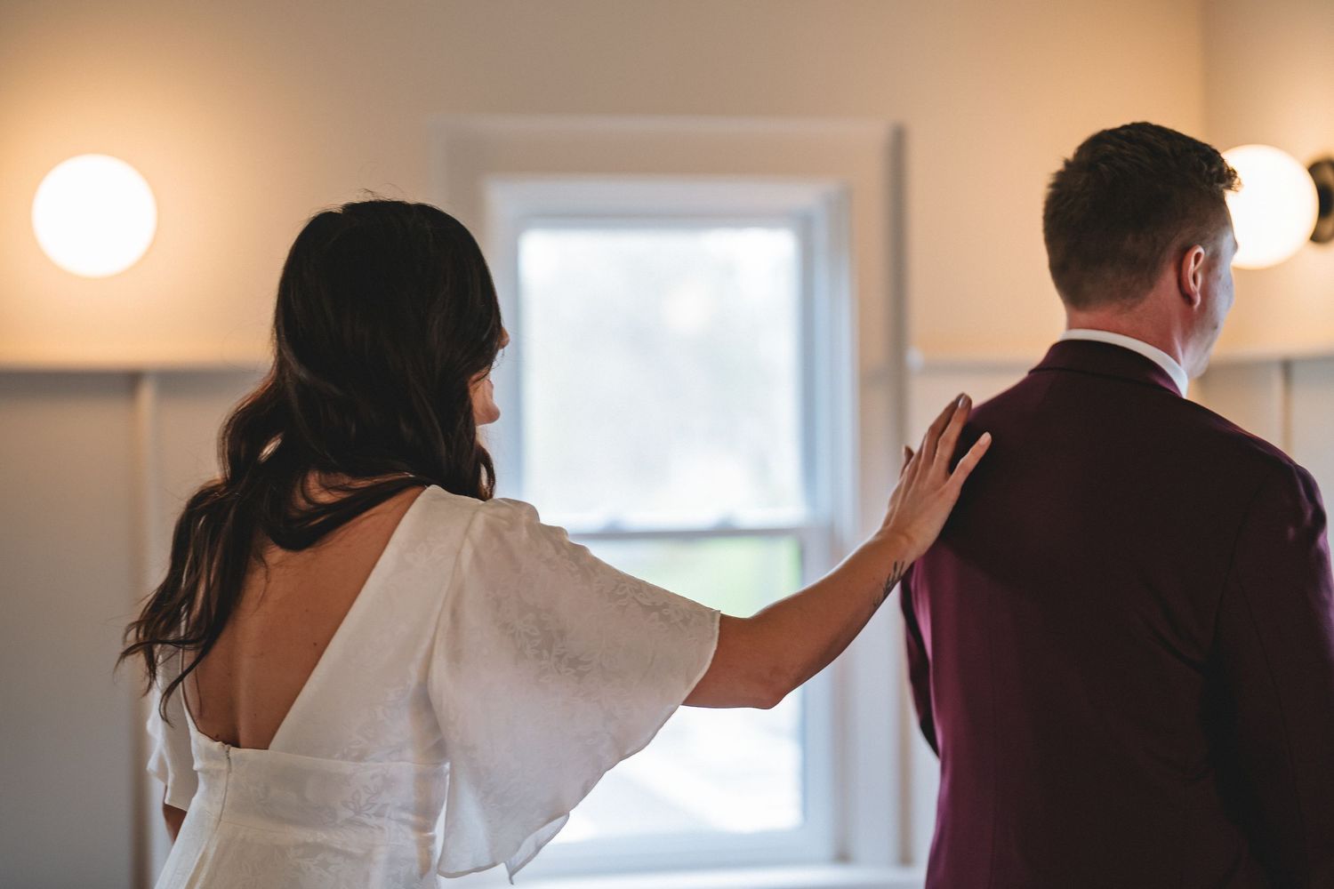 Woman in white dress places hand on companion's shoulder in softly lit indoor setting with window in background.