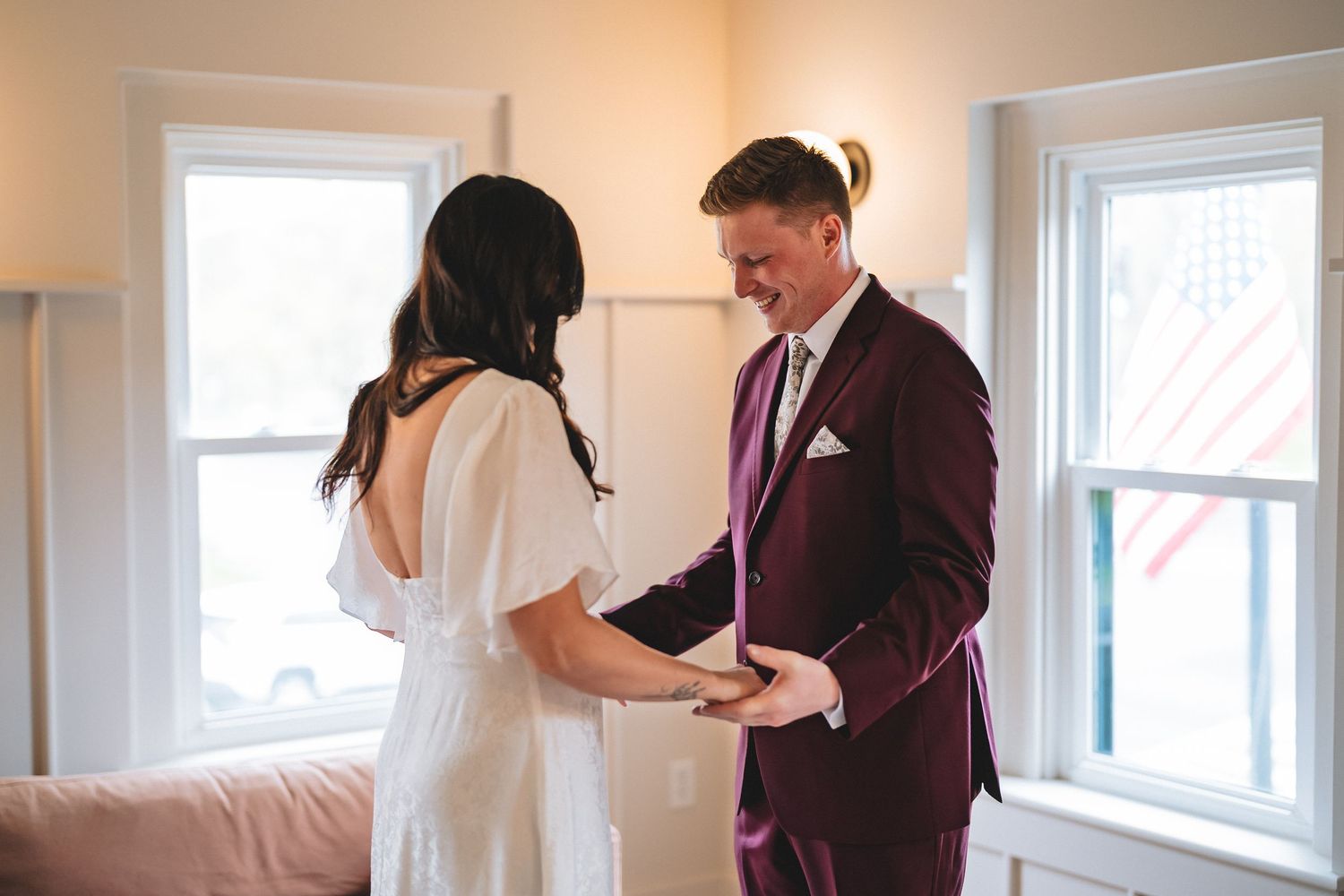 A couple shares a tender first-look moment in a bright sunlit room with white walls and windows.