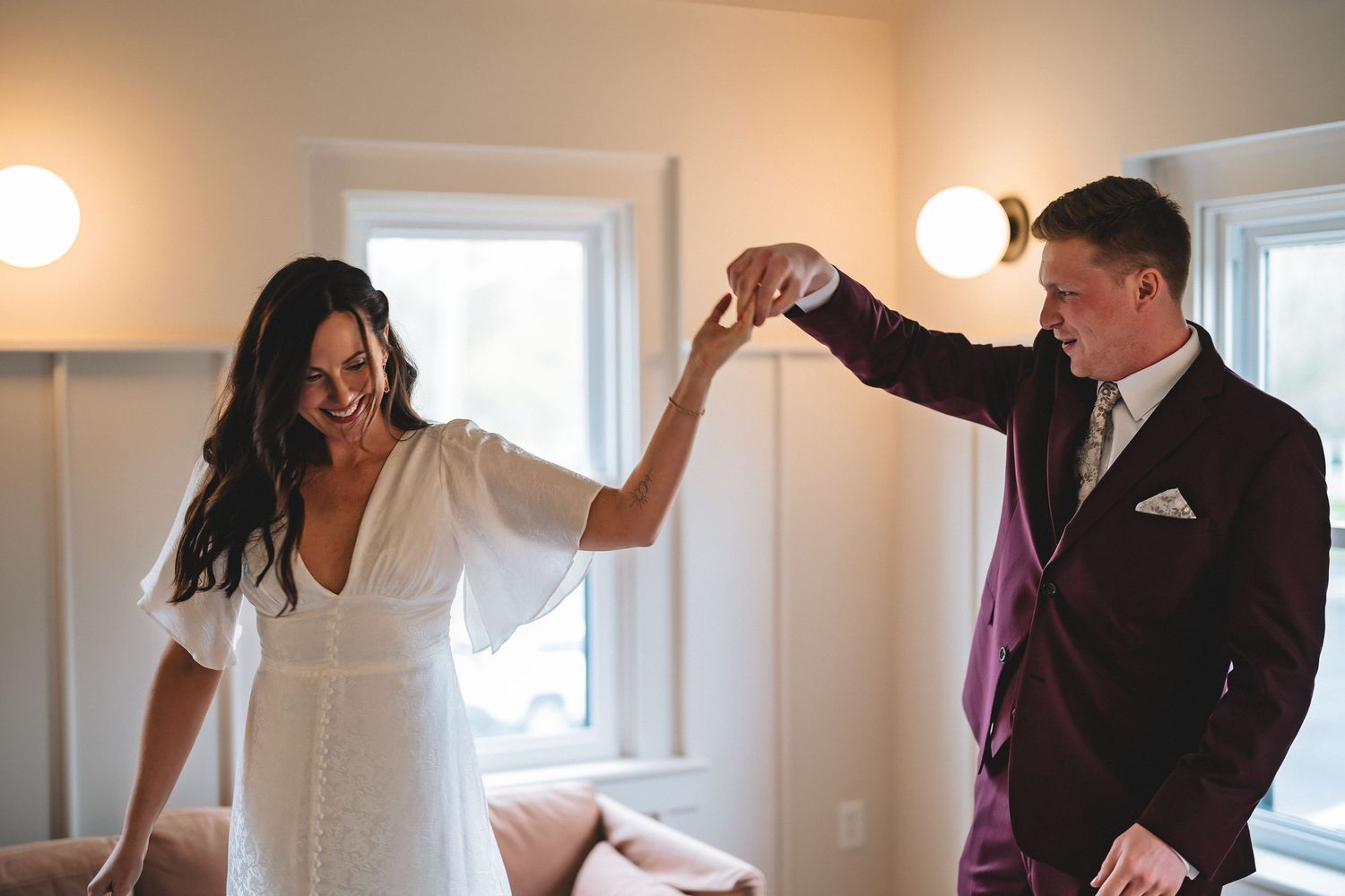 A couple dressed in a white dress and black suit dancing together in a bright room with wall sconces.