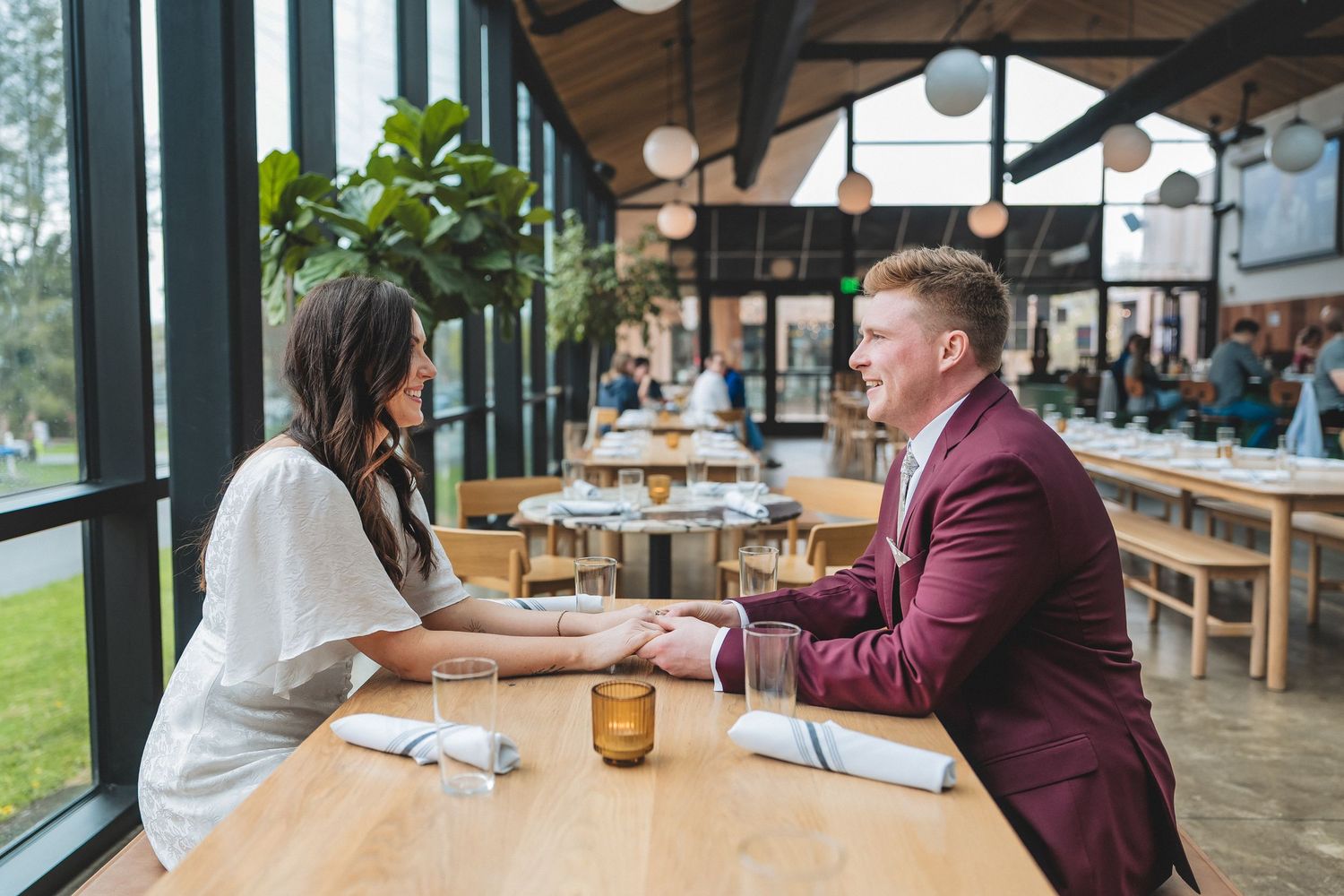 Two people sit facing each other at a wooden table in a modern restaurant with large windows.