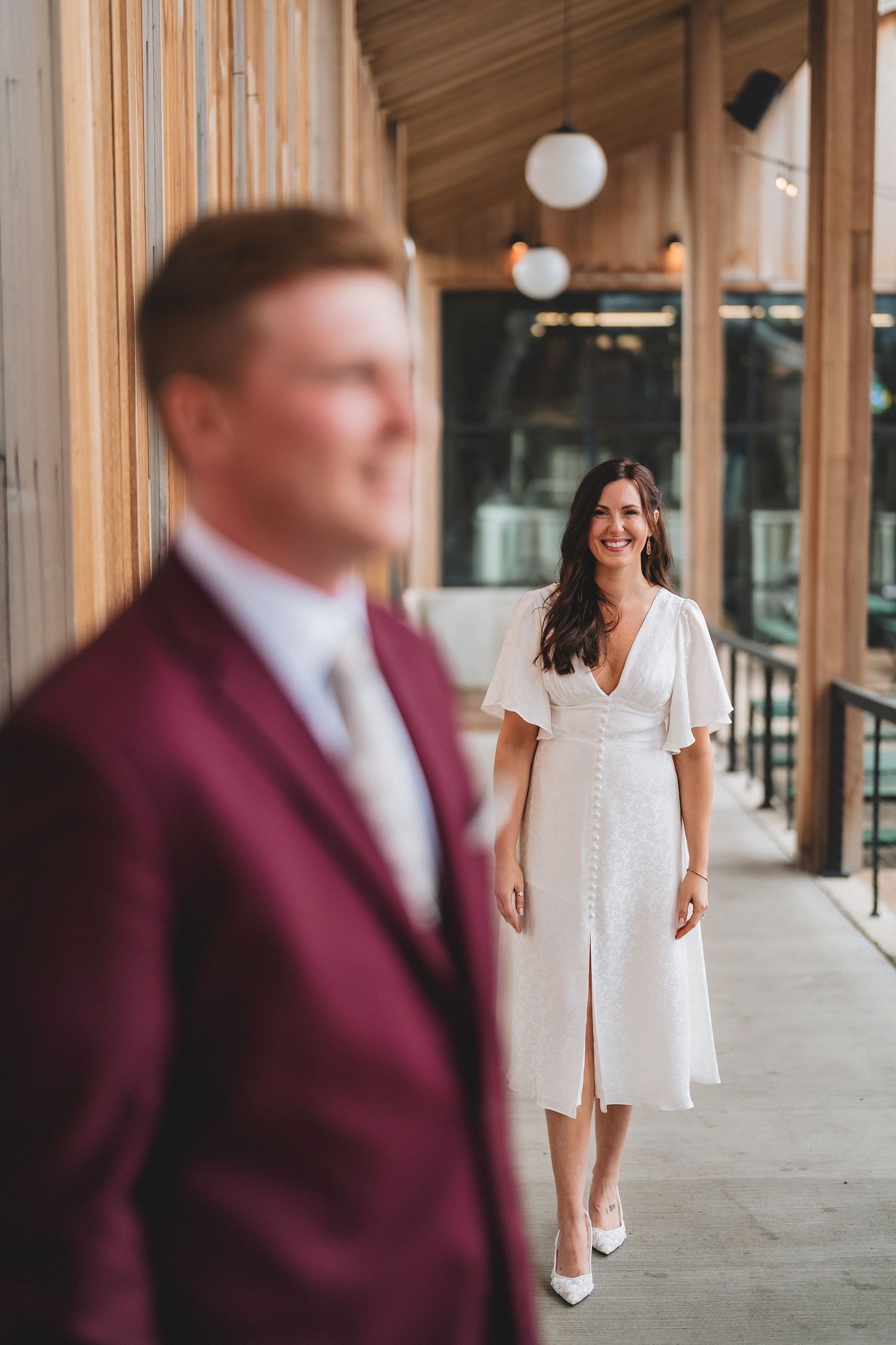 A woman in a white dress smiles while standing on a covered porch as someone in a burgundy suit stands in the foreground.