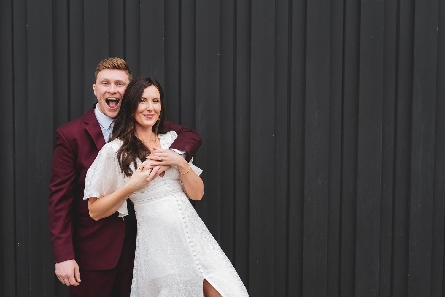A happy couple poses against a dark wall, with the bride in white and groom in burgundy suit.