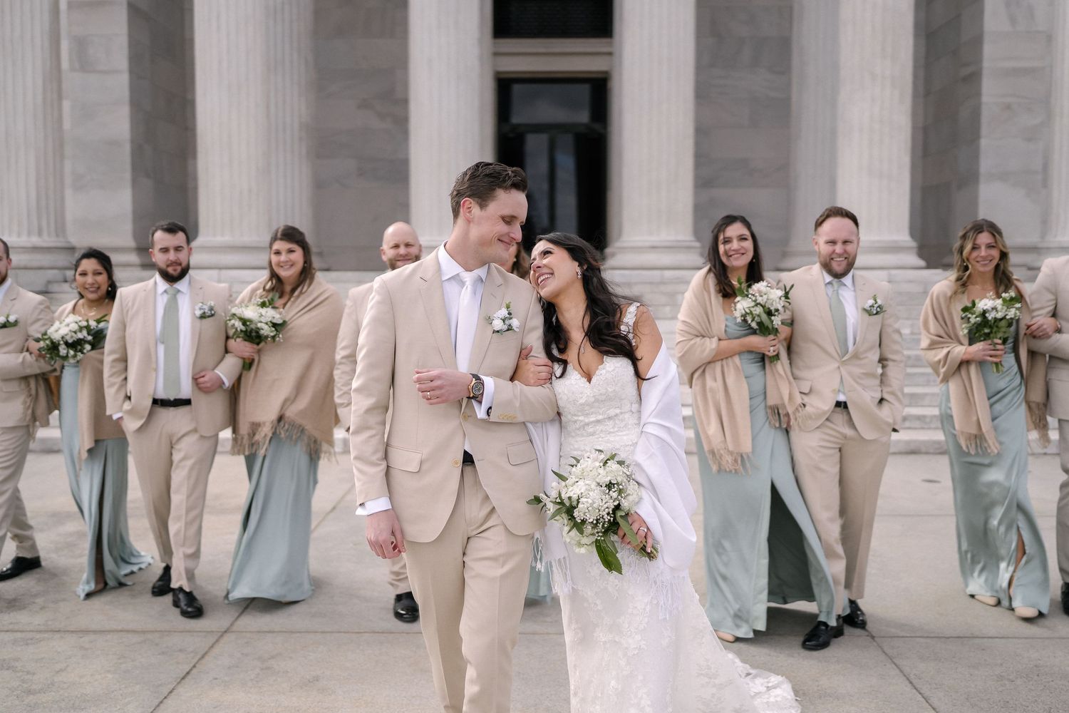 Wedding party in soft neutral tones stands on stone steps as couple walks forward with bouquets and formal attire.
